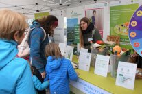 Kinder und Frauen stehen an einem Stand mit Infoftafeln und Lebensmitteln.