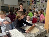 Kinder und die Referentin stehen an einer Arbeitsfläche, auf der ein Kind Teig ausrollt.