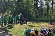 Kinder stehen versammelt auf einer Waldlichtung. Ein Erwachsener erklärt eine Aufgabe.