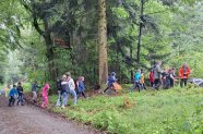 Kinder in Regenkleidung laufen über einen Waldweg zu einem Tisch, hinter dem ein Mann steht