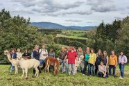 In grüner Hügellandschaft hat sich eine große Gruppe von Frauen und Männern mit vier Alpakas zum Foto zusammengestellt.