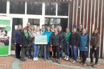 eine Gruppe Menschen vor einer Holz- und Fensterwand. Links davon ein Rollup "Schule geht auf den Bauernhof".