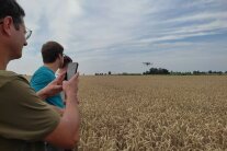 Über einem Weizenfeld fliegt eine Drohne. Am Rand des Feldes beobachten Männer den Vorgang und filmen mit dem Handy.